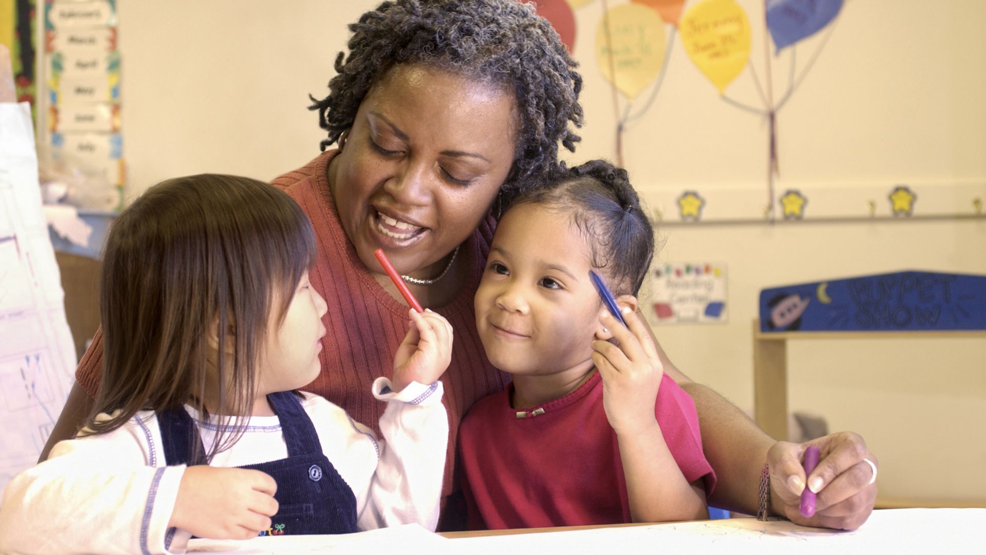 A teacher smiles and works with two small children in an early childhood classroom